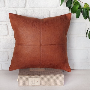 May include: A brown leather pillow with a square stitched design. The pillow is sitting on a book with the title "The Complete Sherlock Holmes" on the spine.