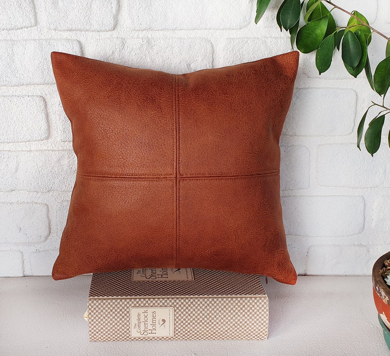 May include: A brown leather pillow with a square stitched design. The pillow is sitting on a book with the title "The Complete Sherlock Holmes" on the spine.