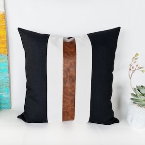 May include: Black and white striped cushion with a brown leather accent strip. The cushion is made of a soft, textured fabric.