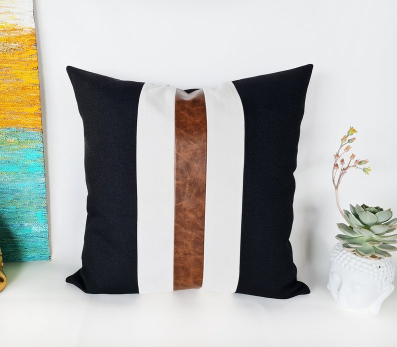 May include: Black and white striped cushion with a brown leather accent strip. The cushion is made of a soft, textured fabric.