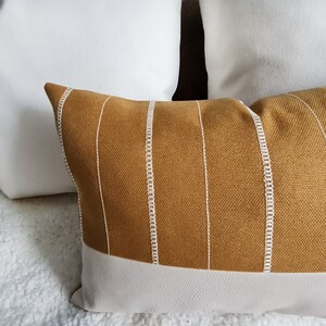 May include: A mustard yellow throw pillow with white piping and a white stripe pattern. The pillow has a white bottom edge.