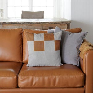 May include: A brown leather sofa with two throw pillows. One pillow is a light gray with brown squares and the other is a solid gray.