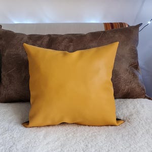 May include: A yellow throw pillow with a brown leather-like pillow behind it. Both pillows are on a white fluffy bed.