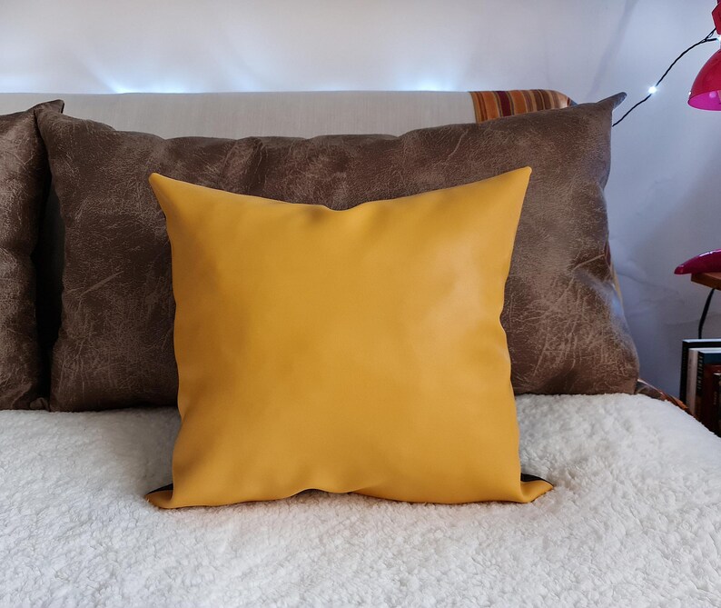 May include: A yellow throw pillow with a brown leather-like pillow behind it. Both pillows are on a white fluffy bed.