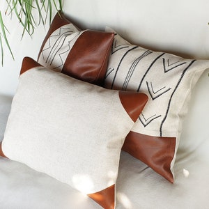 May include: Three decorative throw pillows with a white linen fabric and brown leather accents. The pillows feature geometric patterns in black thread.