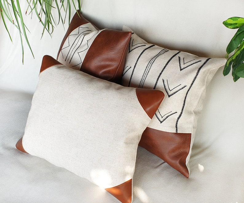 May include: Three decorative throw pillows with a white linen fabric and brown leather accents. The pillows feature geometric patterns in black thread.