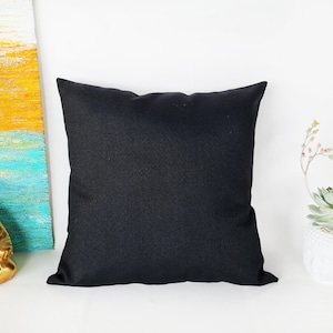 May include: A black square cushion with a smooth, solid surface. The cushion is on a white surface.