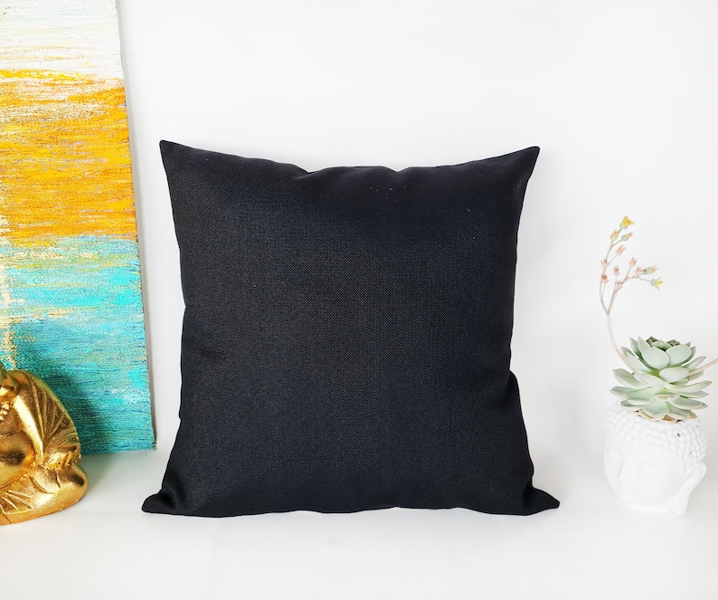 May include: A black square cushion with a smooth, solid surface. The cushion is on a white surface.