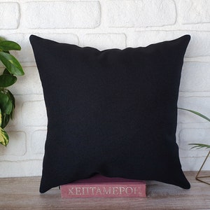 May include: A square black decorative pillow with a textured surface. The pillow is resting on a pink book with the title "HEPTAMERON". The background is a white brick wall with green plants.