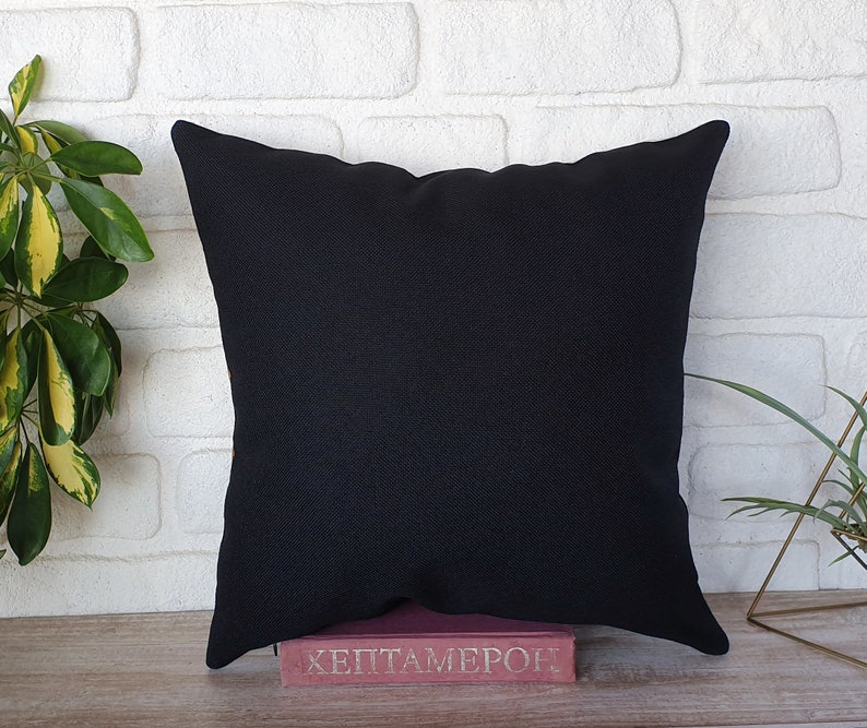 May include: A square black decorative pillow with a textured surface. The pillow is resting on a pink book with the title "HEPTAMERON". The background is a white brick wall with green plants.
