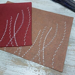 May include: Two placemats with a white embroidered design of stylized leaves on a red and brown background.