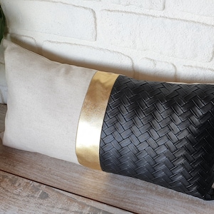 Puede incluir: Una almohada decorativa con un tejido de lino beige en un lado y un tejido de cuero negro tejido en el otro. Una tira dorada separa las dos telas.