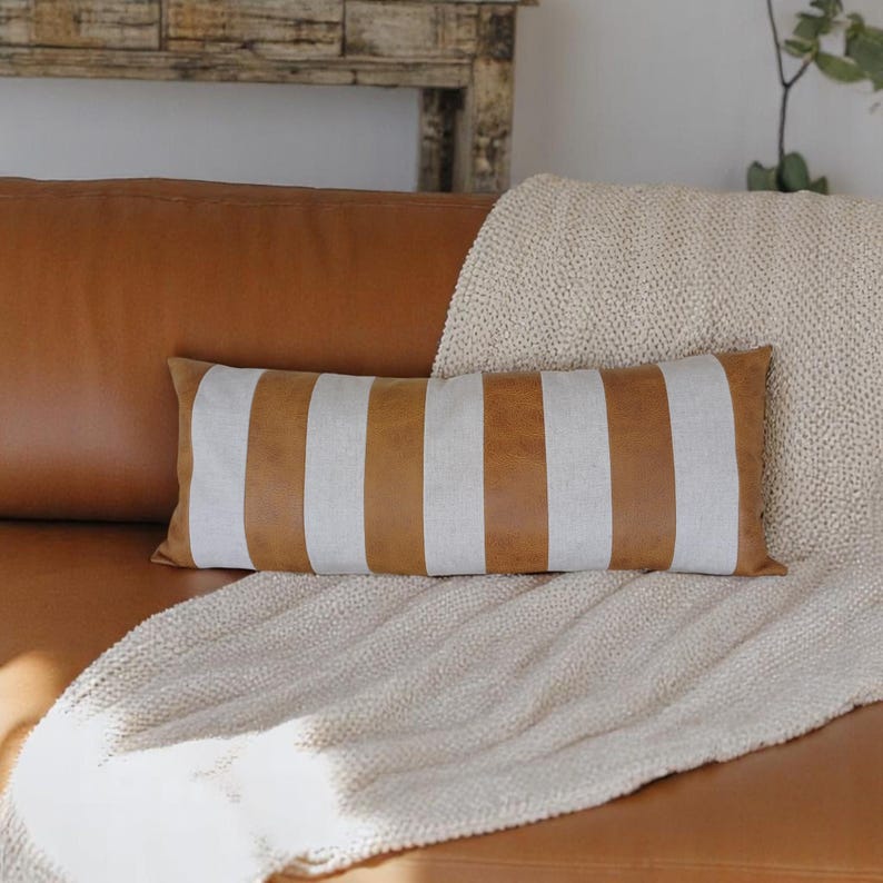 May include: A brown and white striped lumbar pillow with a brown leather look. The pillow is on a light brown couch with a cream throw blanket.