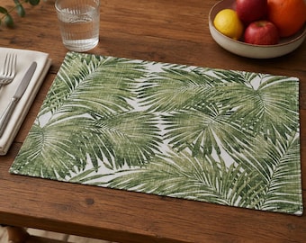 May include: A rectangular placemat featuring a white background with a repeating pattern of green palm leaves. The fabric has a visible woven texture, and the design displays various shades of green, ranging from light to dark, across the leaves.