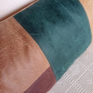May include: A cylindrical bolster cushion featuring a colour-blocked design with panels of textured faux leather in shades of dark teal, tan, and deep brown. The sections are joined by visible stitching, creating a structured, multi-toned appearance.