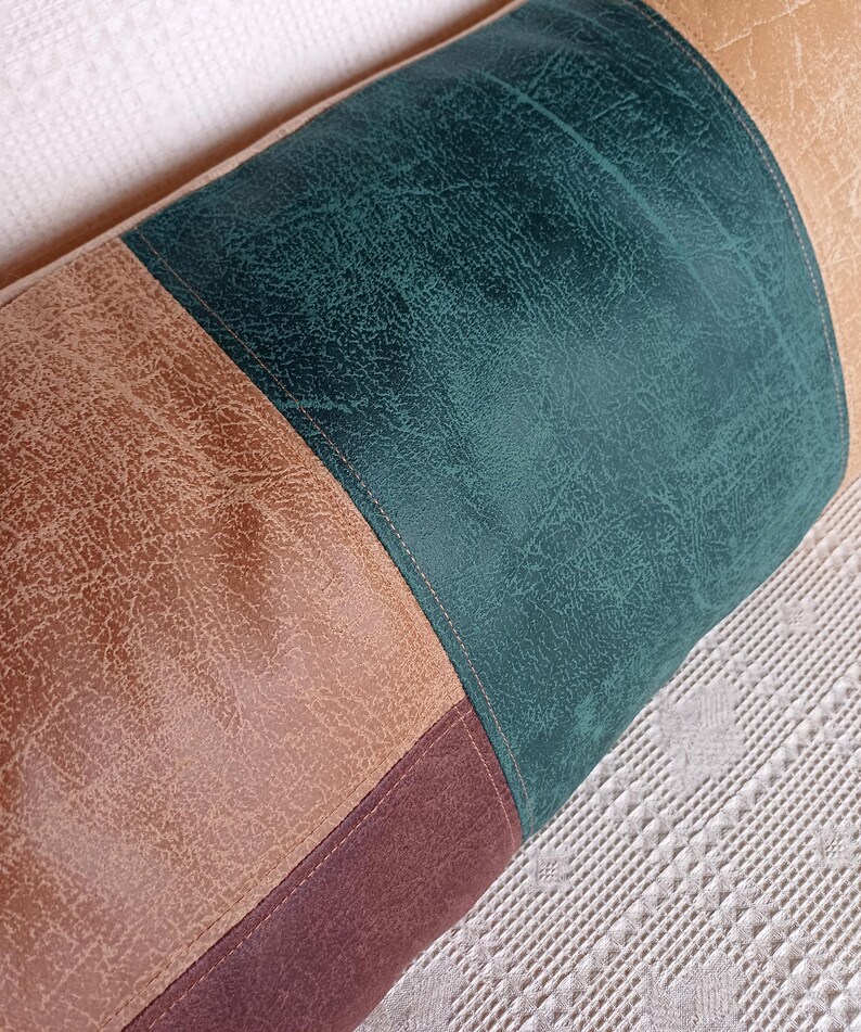 May include: A cylindrical bolster cushion featuring a colour-blocked design with panels of textured faux leather in shades of dark teal, tan, and deep brown. The sections are joined by visible stitching, creating a structured, multi-toned appearance.