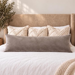 May include: A long, rectangular, solid grey velvet bolster pillow positioned in front of three cream-coloured square throw pillows with a textured, tufted chevron pattern. The pillows are arranged on a white bedspread against a tan headboard.