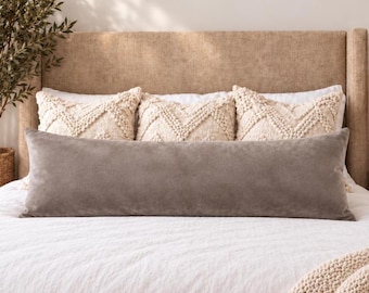 May include: A long, rectangular, solid gray velvet lumbar pillow positioned in front of three square, cream-colored throw pillows with a textured, raised chevron pattern. The pillows are arranged on a white bedspread against a tan headboard.