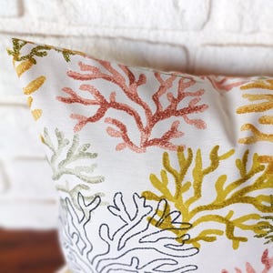 Pu&ograve; includere: Cuscino decorativo bianco con un motivo a barriera corallina nei toni del rosso, giallo, verde e nero. Il cuscino presenta un motivo ripetuto di rami di corallo su sfondo bianco, creando un tema costiero.