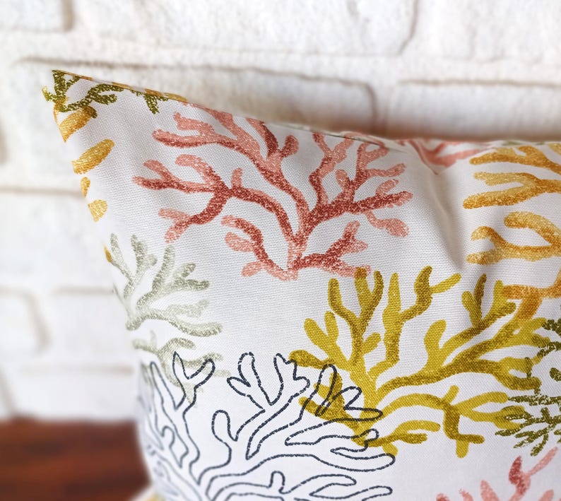 Pu&ograve; includere: Cuscino decorativo bianco con un motivo a barriera corallina nei toni del rosso, giallo, verde e nero. Il cuscino presenta un motivo ripetuto di rami di corallo su sfondo bianco, creando un tema costiero.