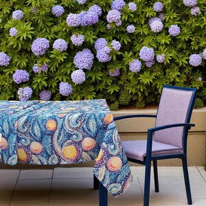 Può includere: Un set da pranzo blu per esterni con una tovaglia fantasia e una sedia abbinata. La tovaglia presenta un design a vortice con accenti gialli e rosa. La sedia ha seduta e schienale lavanda, con una struttura blu scuro. Lo sfondo è una siepe verde con fiori viola.