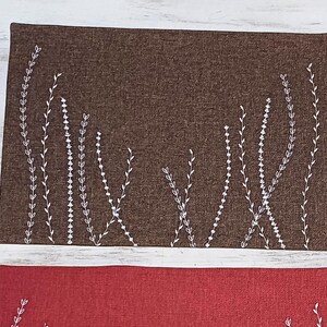 May include: Two placemats with a white embroidered floral design on a brown and red fabric background.