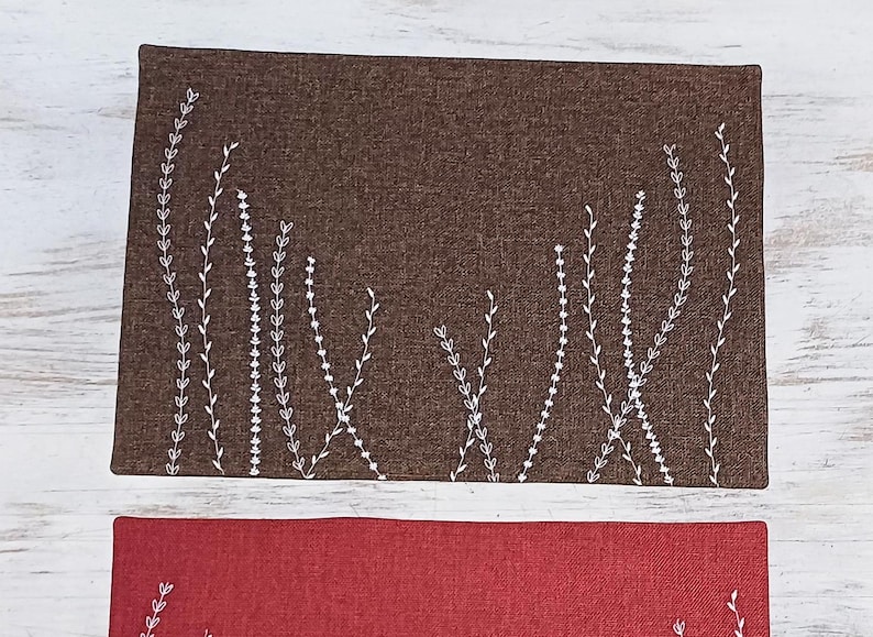 May include: Two placemats with a white embroidered floral design on a brown and red fabric background.