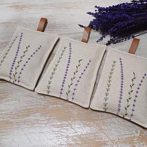 May include: Three linen lavender sachets with embroidered lavender sprigs in shades of purple and green. The sachets have brown leather ties.