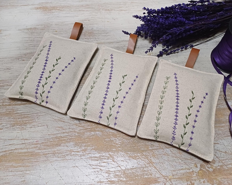 May include: Three linen lavender sachets with embroidered lavender sprigs in shades of purple and green. The sachets have brown leather ties.