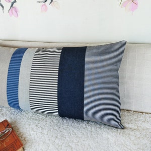 May include: A long, rectangular pillow with a striped pattern. The pillow is made of different fabrics in shades of blue, white, and black. The pillow is lying on a white, fluffy surface.