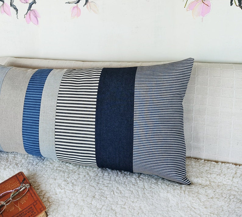 May include: A long, rectangular pillow with a striped pattern. The pillow is made of different fabrics in shades of blue, white, and black. The pillow is lying on a white, fluffy surface.