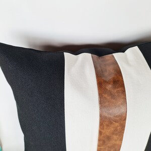 May include: Black and white striped cushion with a brown leather accent stripe. The cushion is made of a soft, textured fabric.