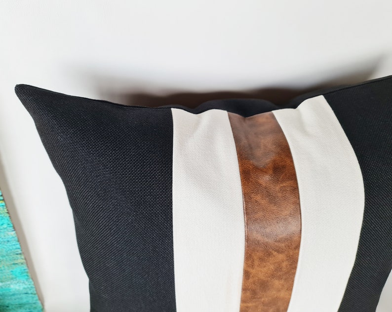 May include: Black and white striped cushion with a brown leather accent stripe. The cushion is made of a soft, textured fabric.