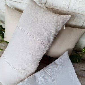 May include: Three decorative throw pillows with a textured, faux leather fabric. The pillows are in shades of cream and white.