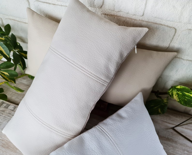 May include: Three decorative throw pillows with a textured, faux leather fabric. The pillows are in shades of cream and white.