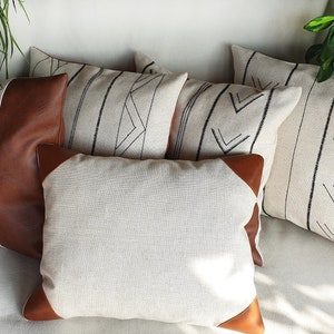 May include: Five decorative throw pillows with geometric patterns. The pillows are made of a light beige linen fabric with black embroidery. Two of the pillows have brown leather accents.