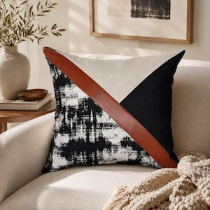 May include: Decorative square pillow with a geometric design. The pillow features a black and white abstract pattern, a solid black triangle, and a beige triangle. A brown leather strap accents the design. The pillow is on a cream-colored chair.