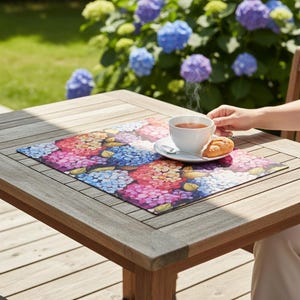 Puede incluir: Una mesa de madera con un mantel individual floral con un vibrante diseño de hortensias en tonos rosa, morado, azul y amarillo. Una taza y un platillo blancos con un cruasán están sobre el mantel. El fondo muestra un jardín con hortensias floreciendo.