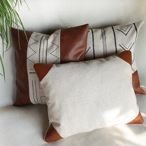 May include: Three decorative throw pillows with geometric patterns. The pillows are made of a beige linen fabric with brown leather accents. The patterns are black and white.