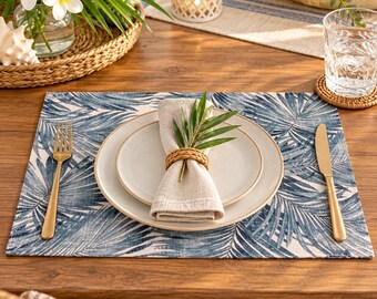 May include: A rectangular placemat featuring a blue and white palm leaf pattern, topped with a white ceramic plate, a beige linen napkin held by a woven rattan napkin ring, a gold-toned fork on the left, and a gold-toned knife on the right.