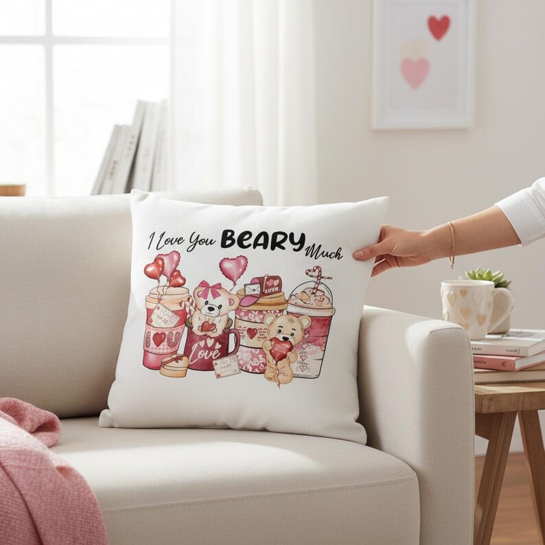 May include: White square pillow with the phrase "I Love You Beary Much" and illustrations of teddy bears and coffee cups. The bears hold hearts and are surrounded by heart-shaped balloons. The pillow is on a white couch.