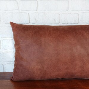 May include: A rectangular, brown leather pillow with a textured surface. The pillow is placed on a wooden surface, with a white brick wall in the background. The pillow is a warm, earthy tone.