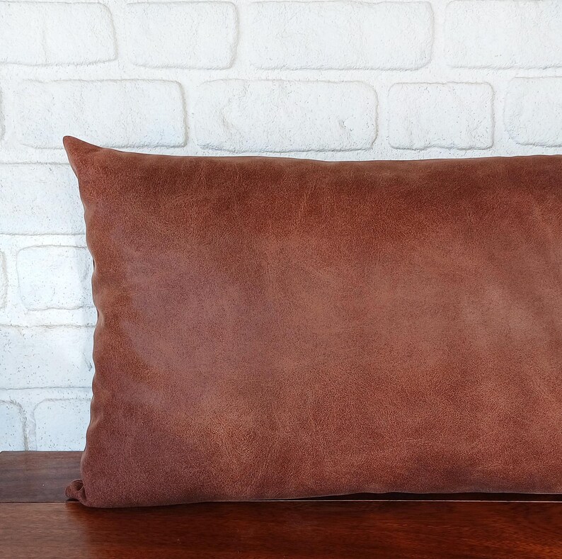 May include: A rectangular, brown leather pillow with a textured surface. The pillow is placed on a wooden surface, with a white brick wall in the background. The pillow is a warm, earthy tone.