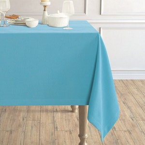 May include: A light blue tablecloth with a smooth, even texture. The tablecloth is draped over a wooden table with turned legs. The tablecloth is set with a white bowl, a white serving dish, and two clear wine glasses.