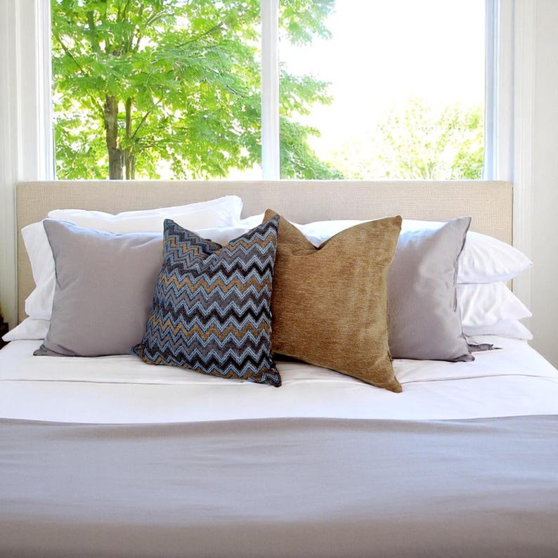 Op de afbeelding: Een bed met een neutraal gekleurd hoofdbord en wit beddengoed. Vier decoratieve kussens in grijstinten, bruin en een chevronpatroon zijn erop geplaatst. Op de achtergrond een groot raam met uitzicht op een boom.