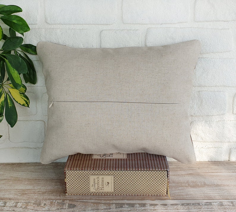 May include: A beige linen pillow with a zipper closure. The pillow is sitting on a book with a brown and white checkered pattern. The book has the text "The Complete Sherlock Holmes" on the spine.
