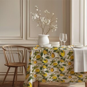 May include: A dining table set with a white tablecloth and a lemon-patterned tablecloth. The table is set with white dishes, wine glasses, and a white pitcher with white flowers. A wooden chair is on the left.