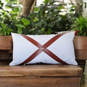 Könnte beinhalten: Ein rechteckiges weißes Kissen mit braunen Lederriemen, die ein X über die Vorderseite bilden und mit einer silbernen Niete befestigt sind. Das Kissen liegt auf einer Holzbank, mit Grünzeug im Hintergrund.