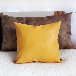 May include: A yellow throw pillow with a brown leather pillow behind it. Both pillows are on a white fluffy surface.