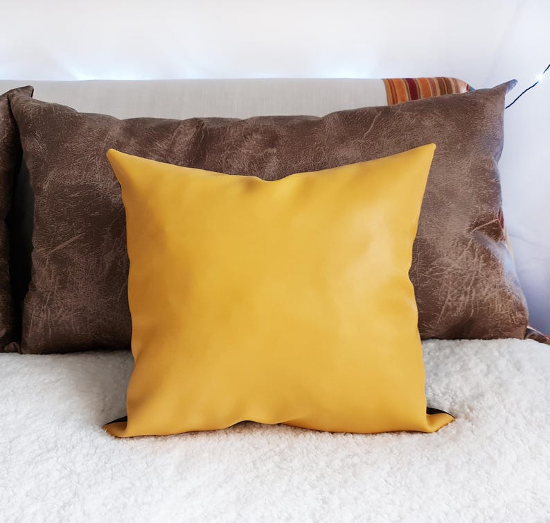 May include: A yellow throw pillow with a brown leather pillow behind it. Both pillows are on a white fluffy surface.